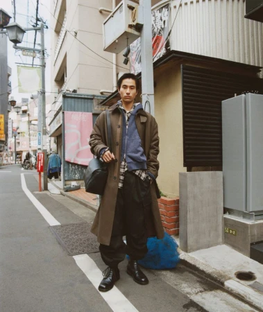 Man wearing a brown trench coat and black trousers standing on a narrow city street holding a shoulder bag