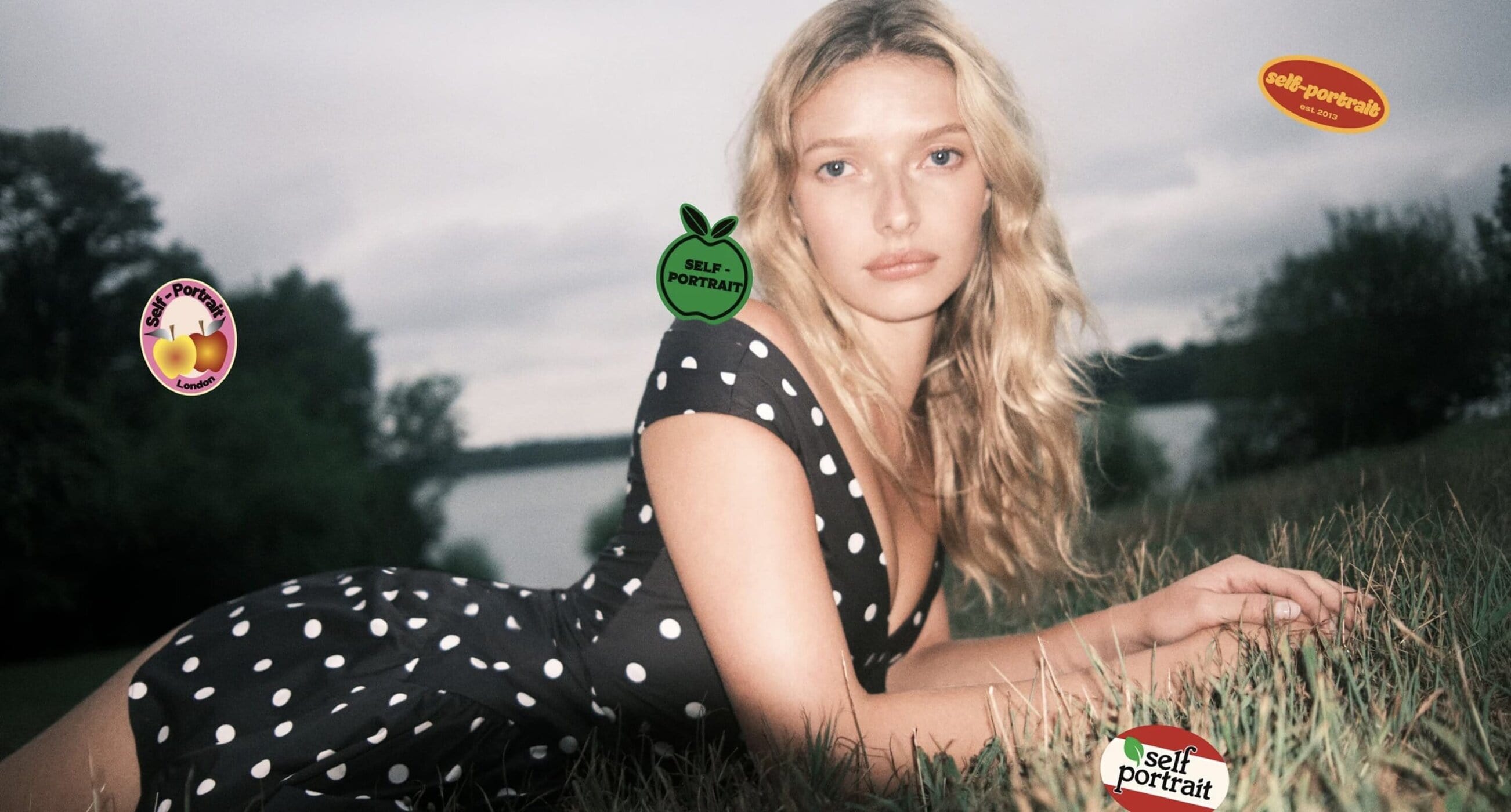 Blonde woman in a polka dot dress lies in a grassy field near a body of water, with stickers of the brand logo floating around.
