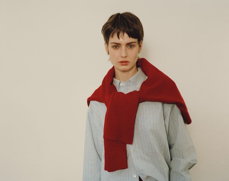 Young woman in a striped shirt with a red sweater draped over her shoulders posing against a white background indoors with soft lighting.