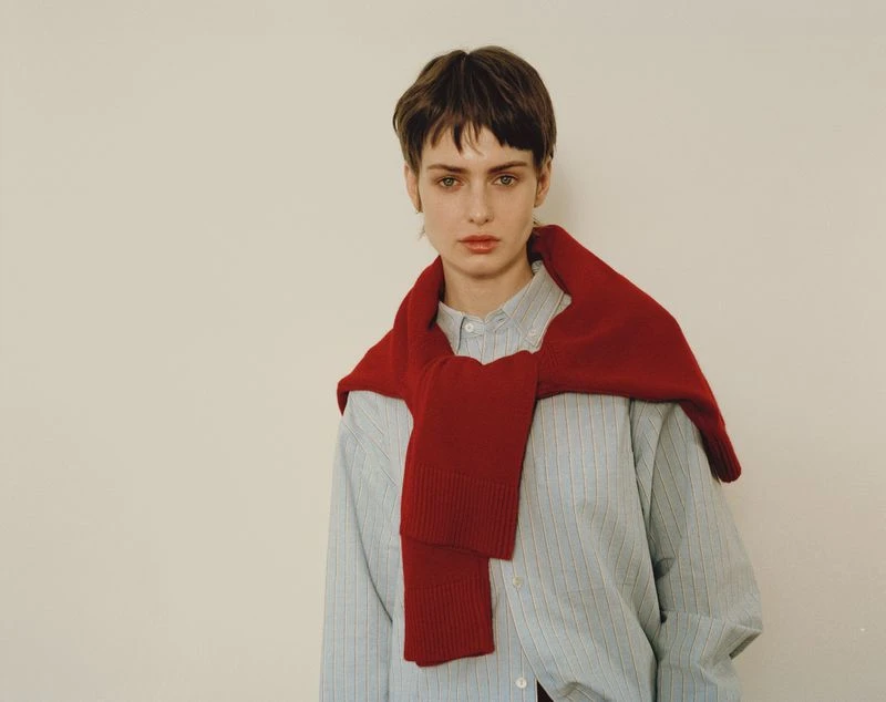 Young woman in a striped shirt with a red sweater draped over her shoulders posing against a white background indoors with soft lighting.