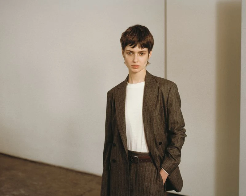 A stylish adult female model poses indoors wearing a brown pinstripe suit, a white t-shirt, and a brown belt against a neutral background.
