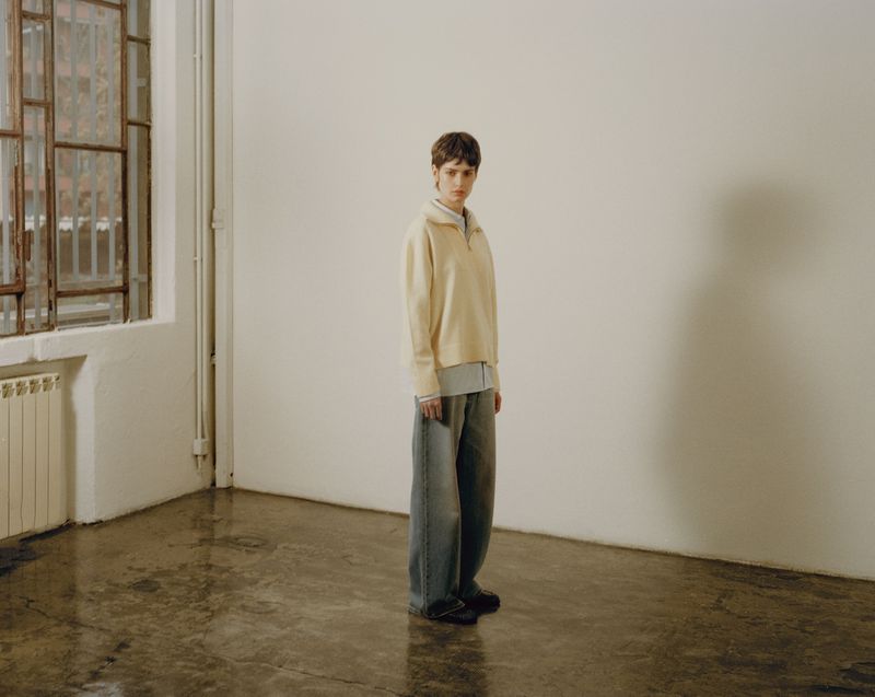 Indoor fashion shot featuring a young adult with short brunette hair wearing a cream top and blue denim baggy pants in a studio.