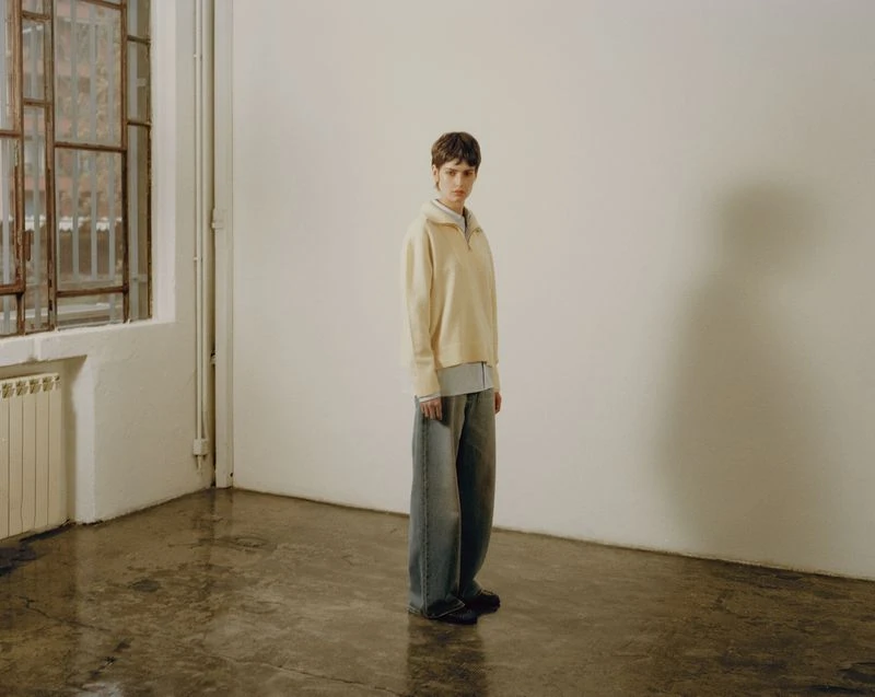Indoor fashion shot featuring a young adult with short brunette hair wearing a cream top and blue denim baggy pants in a studio.