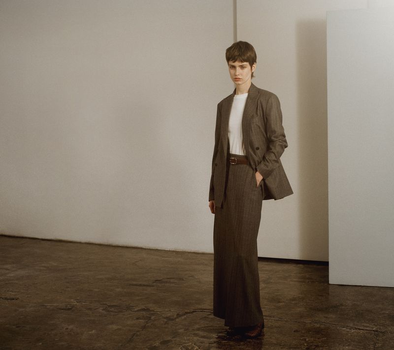 Model standing in a brown blazer and long skirt against a minimalist wall