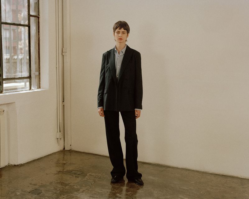 Model standing in a black blazer and trousers against a plain white wall with a window to the left.