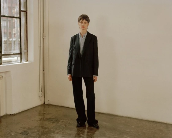 Model standing in a black blazer and trousers against a plain white wall with a window to the left.