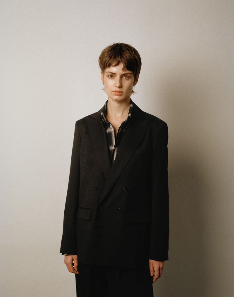 Model with short brown hair wearing a black double-breasted blazer standing against a plain neutral background.