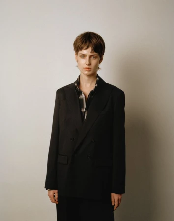 Model with short brown hair wearing a black double-breasted blazer standing against a plain neutral background.