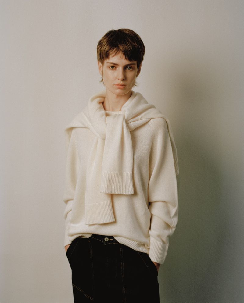 Fashion shot of a female model in a white sweater and black pants against a white backdrop, posing with hands in her pockets.