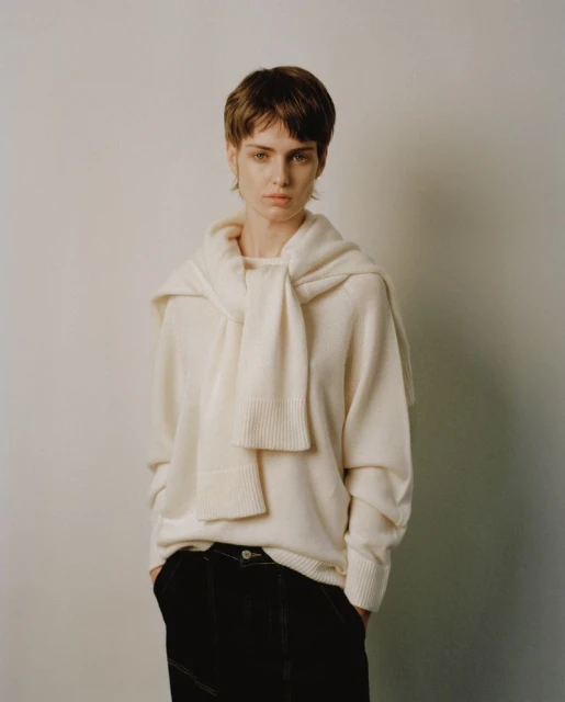 Fashion shot of a female model in a white sweater and black pants against a white backdrop, posing with hands in her pockets.