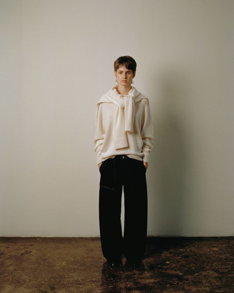 A woman stands indoors wearing a cream sweater, a sweater tied around her shoulders, black pants, and black shoes against a wall.