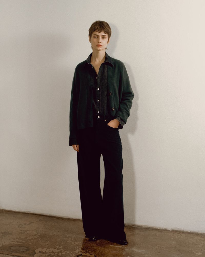 A woman in a jacket and jeans stands against a white wall. The clothing is casual and the lighting is soft and diffused.