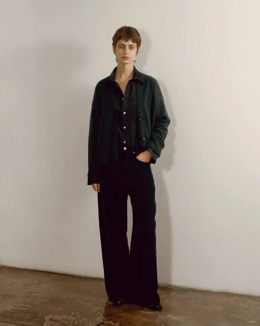 A woman in a jacket and jeans stands against a white wall. The clothing is casual and the lighting is soft and diffused.
