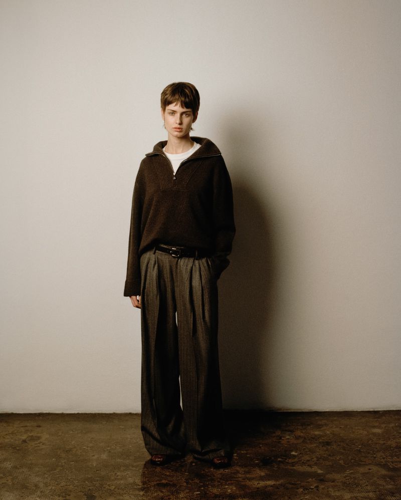 A young adult woman poses in a studio wearing a dark brown pullover sweater, wide leg grey trousers, and a black belt against a neutral backdrop.