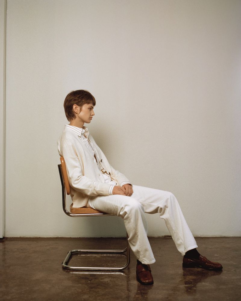 Adult with short hair sits on a chrome chair in front of a white wall, wearing white button-down shirt and pants with brown leather dress shoes.