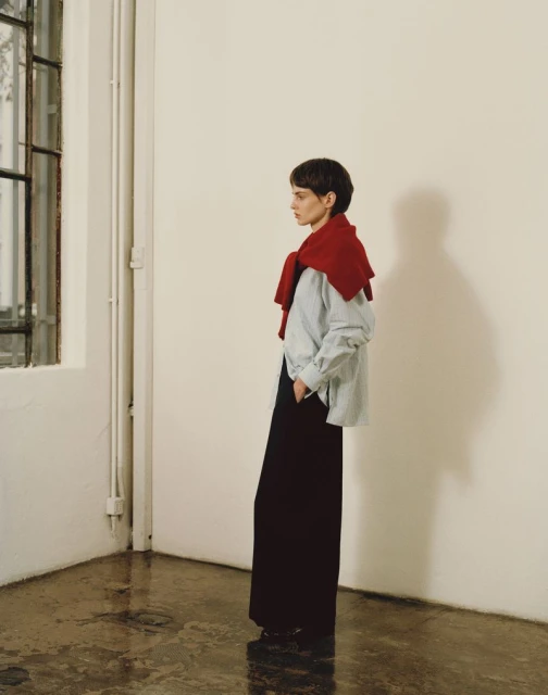 A woman with short hair, wearing wide-leg pants and a light blue shirt with a red sweater draped over her shoulders, poses indoors.