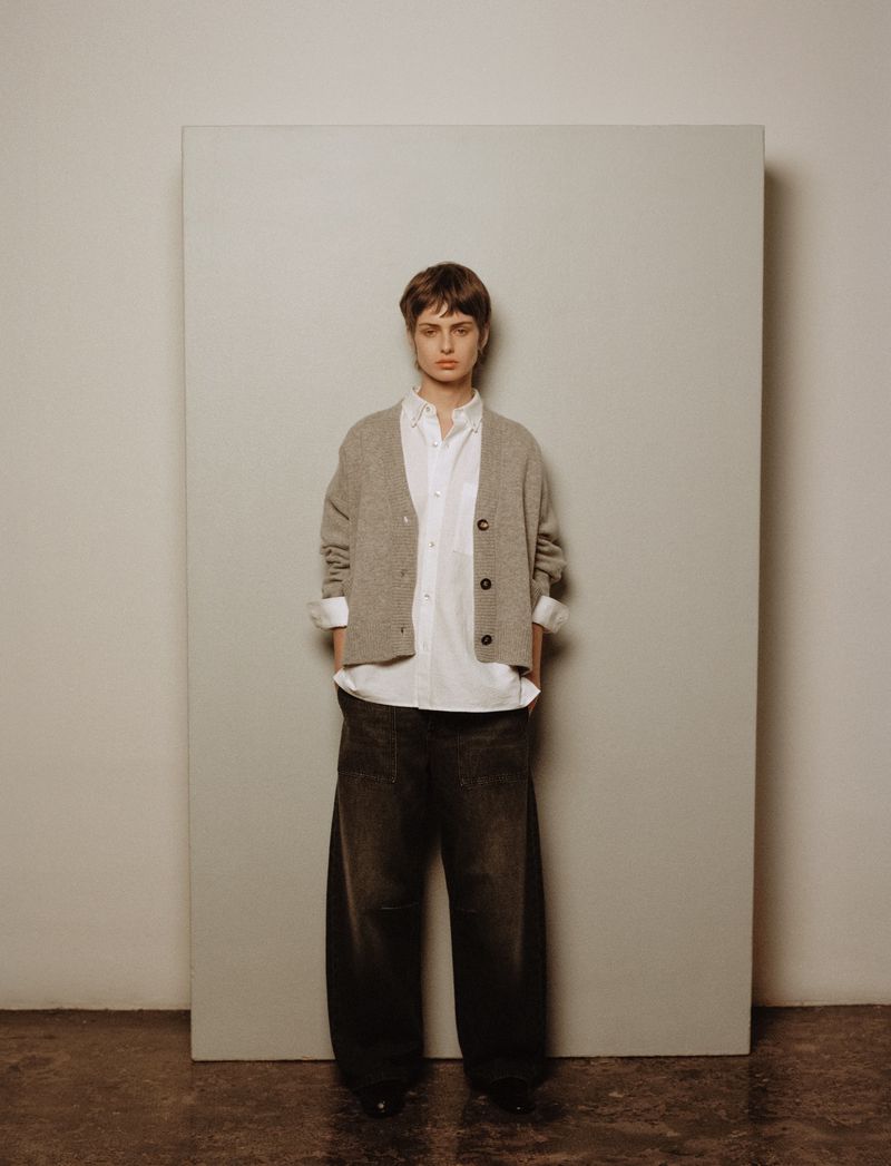 Model wearing a grey cardigan over a white button-down shirt and dark trousers against a neutral panel backdrop.