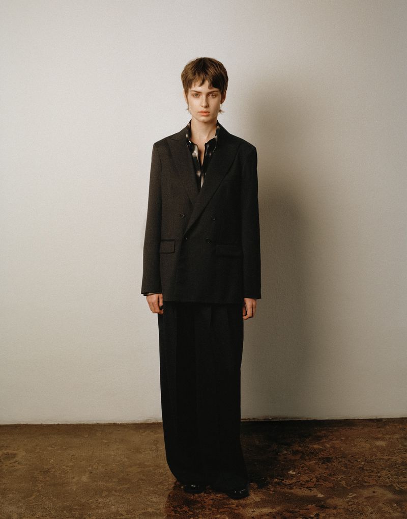 Model standing against a neutral wall wearing a dark blazer and matching trousers for Anaii F/W 2026 Lookbook