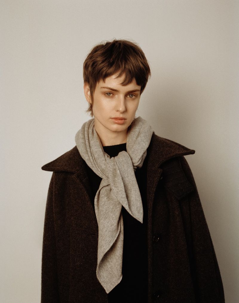 Portrait of a woman wearing a grey scarf and a brown coat, posing in front of a neutral background in a fashion-style photograph.