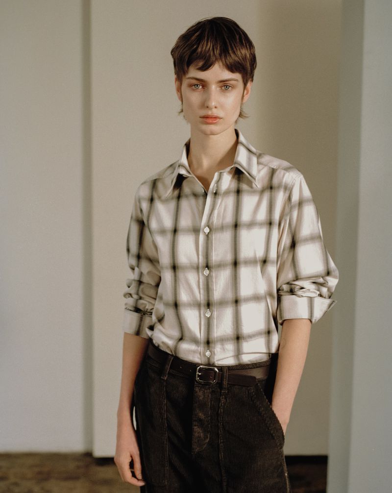 Indoor portrait of a young adult woman with short brown hair. She is wearing a plaid button-down shirt and corduroy pants.
