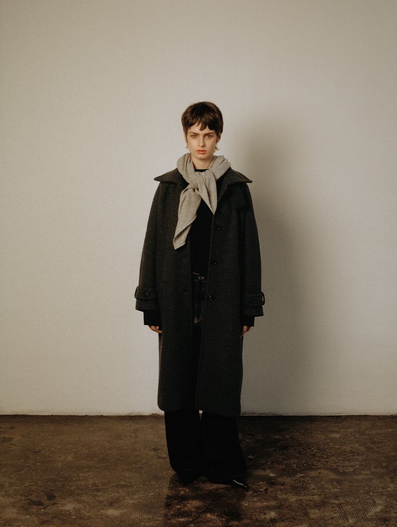 Indoor studio portrait of a female adult with short dark hair wearing a long coat and light-grey scarf against an off-white background.