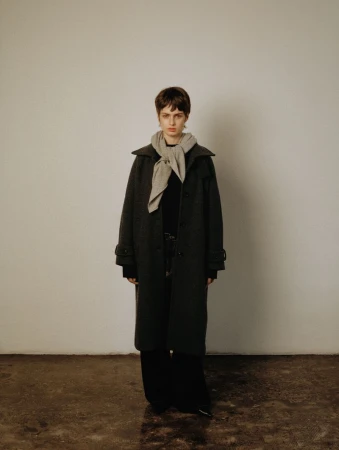 Indoor studio portrait of a female adult with short dark hair wearing a long coat and light-grey scarf against an off-white background.