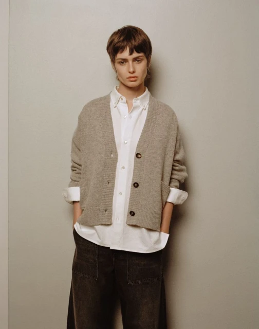Woman in a white button-down shirt and grey cardigan stands in front of a grey wall, hands in her pockets, looking at the camera.