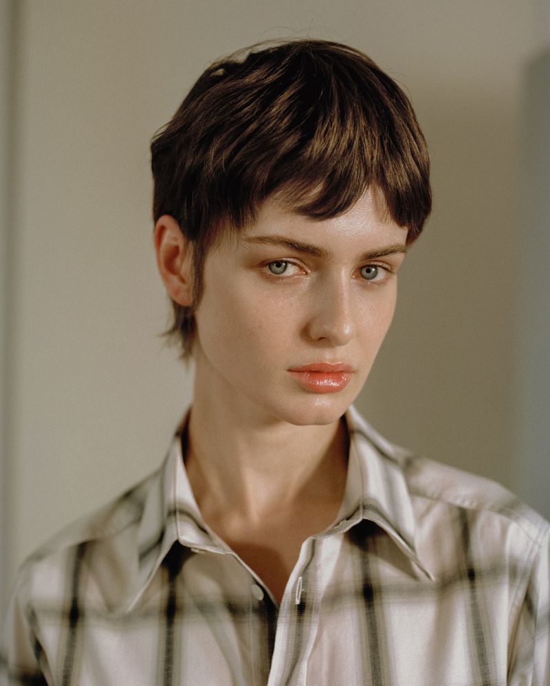 Close-up portrait of a model with short brown hair wearing a plaid button-down shirt against a neutral background.