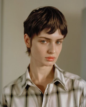Close-up portrait of a model with short brown hair wearing a plaid button-down shirt against a neutral background.