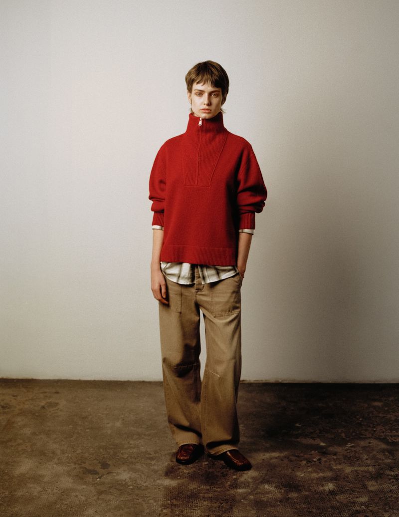 Model in a red high-neck zip sweater and brown trousers standing against a pale grey studio background