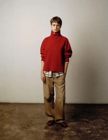 Model in a red high-neck zip sweater and brown trousers standing against a pale grey studio background