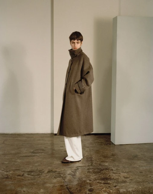 A person in a long brown coat and white pants stands in a minimalist studio with neutral walls and a concrete floor, fashion photography.