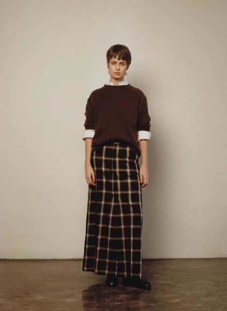 Woman in brown sweater and plaid skirt stands indoors against a neutral wall. Fashion shot with natural lighting and neutral tones.