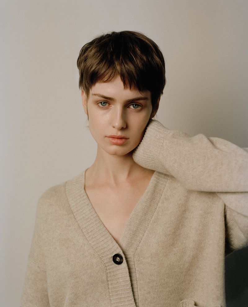 Portrait of a model with short brown hair wearing a beige V-neck cardigan against a plain neutral background.