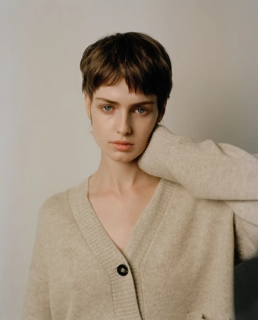 Portrait of a model with short brown hair wearing a beige V-neck cardigan against a plain neutral background.