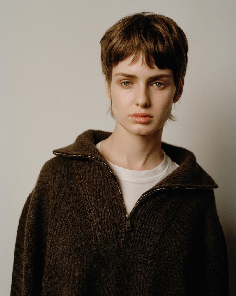 Portrait of a woman with short brown hair wearing a brown zip-up sweater against a plain white background in a studio setting.