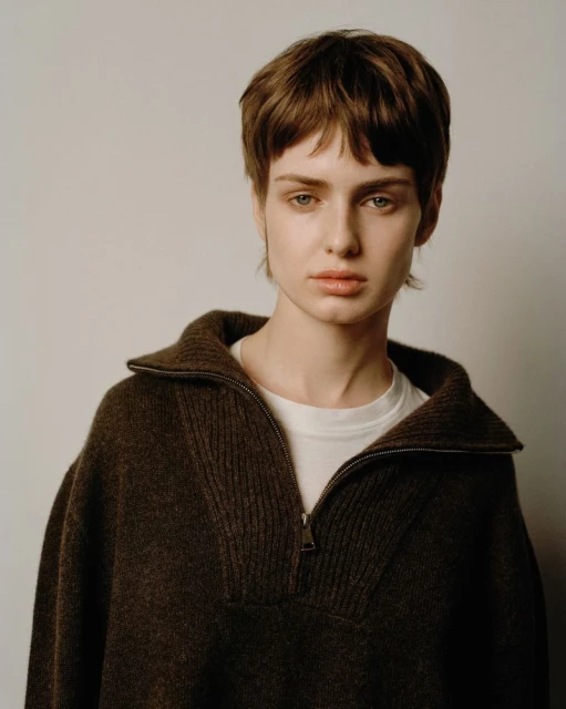 Portrait of a woman with short brown hair wearing a brown zip-up sweater against a plain white background in a studio setting.