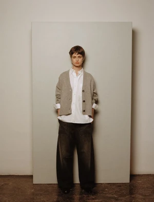 Model wearing a grey cardigan over a white button-down shirt and dark trousers against a neutral panel backdrop.