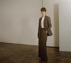 Model standing in a brown blazer and long skirt against a minimalist wall
