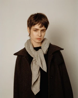 Model with short brown hair wearing a dark coat and light grey scarf against a neutral background