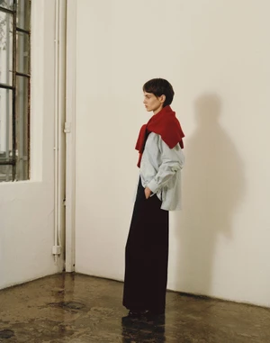 Model standing in profile wearing a light blue buttoned shirt, dark trousers, and a red sweater draped over shoulders.