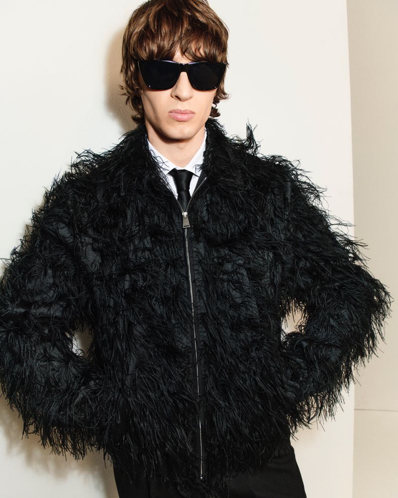 A slender male model with sunglasses poses in a black feather jacket, white shirt, and tie against a plain background in a portrait shot.