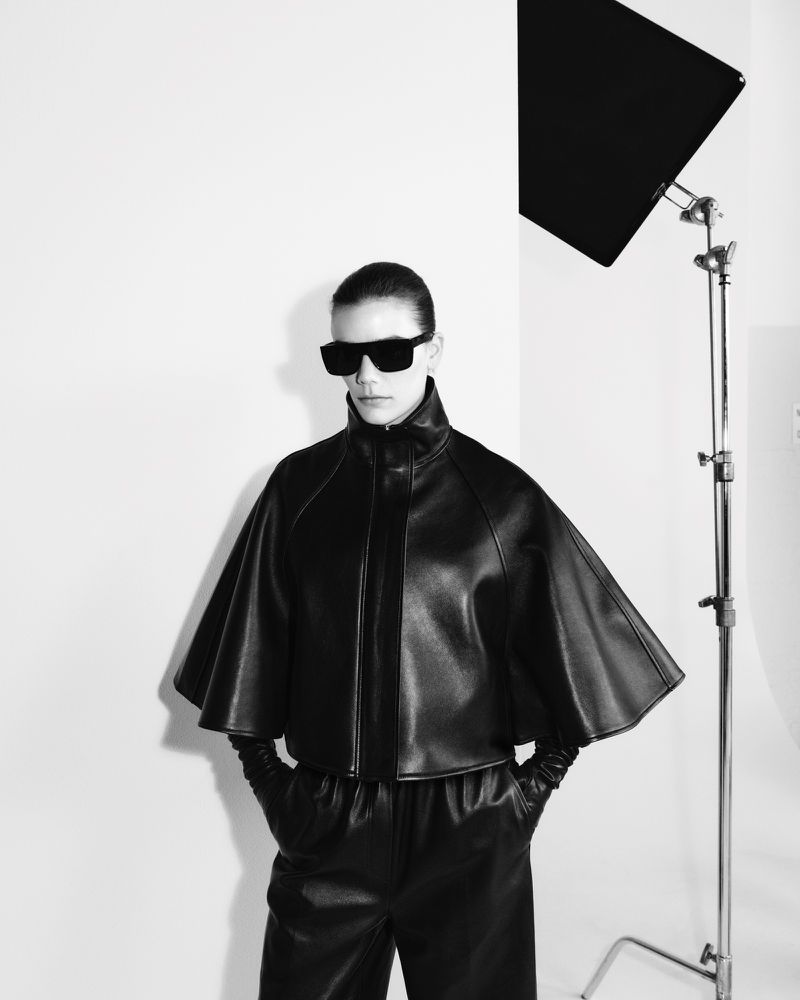 A woman in sunglasses, a black leather jacket with cape-like sleeves, and matching pants stands in a photo studio, illuminated by studio lighting.