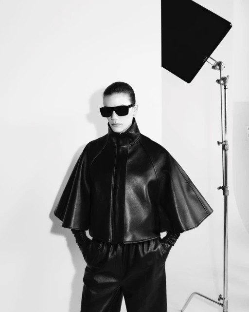 A woman in sunglasses, a black leather jacket with cape-like sleeves, and matching pants stands in a photo studio, illuminated by studio lighting.
