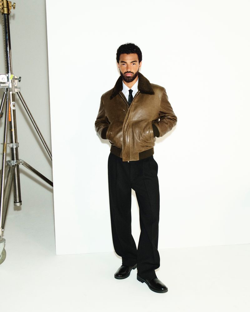 Full-length shot of a man in a brown leather jacket, black pants, and dress shoes, standing in a photo studio with white backdrop.