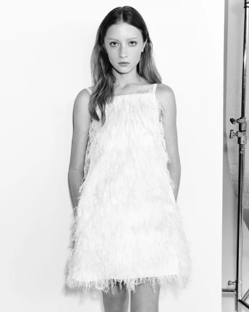 A full shot in black and white shows a young woman in a feathered mini dress, standing in front of a white background in a photo studio.