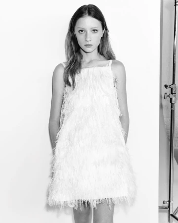 A full shot in black and white shows a young woman in a feathered mini dress, standing in front of a white background in a photo studio.