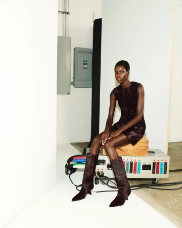 An African-American fashion model sits with tall brown boots and a brown dress in a studio with a large, vintage electrical device.