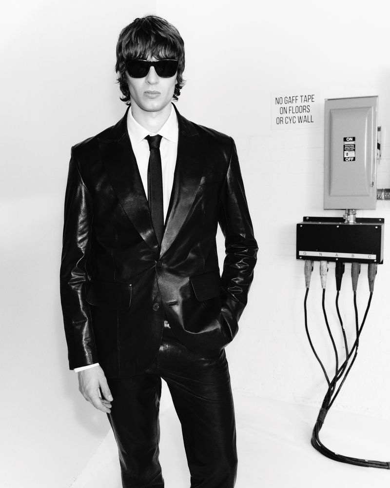 A man in a leather suit and sunglasses stands against a white wall with electrical boxes, creating a bold, monochromatic fashion statement.