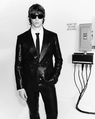 A man in a leather suit and sunglasses stands against a white wall with electrical boxes, creating a bold, monochromatic fashion statement.